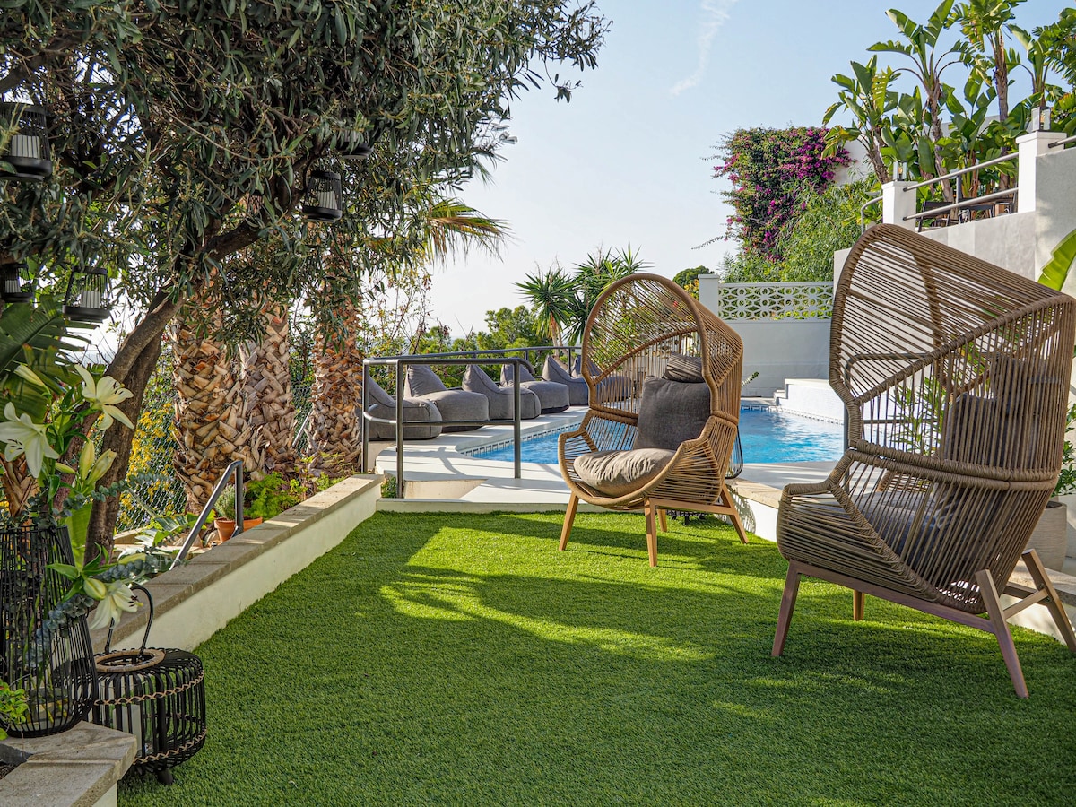A private terrace is surrounded by lush greenery and features two woven lounge chairs. Nestled on a vibrant artificial lawn, the area offers views of a swimming pool and tropical plants, creating a serene outdoor space for relaxation.