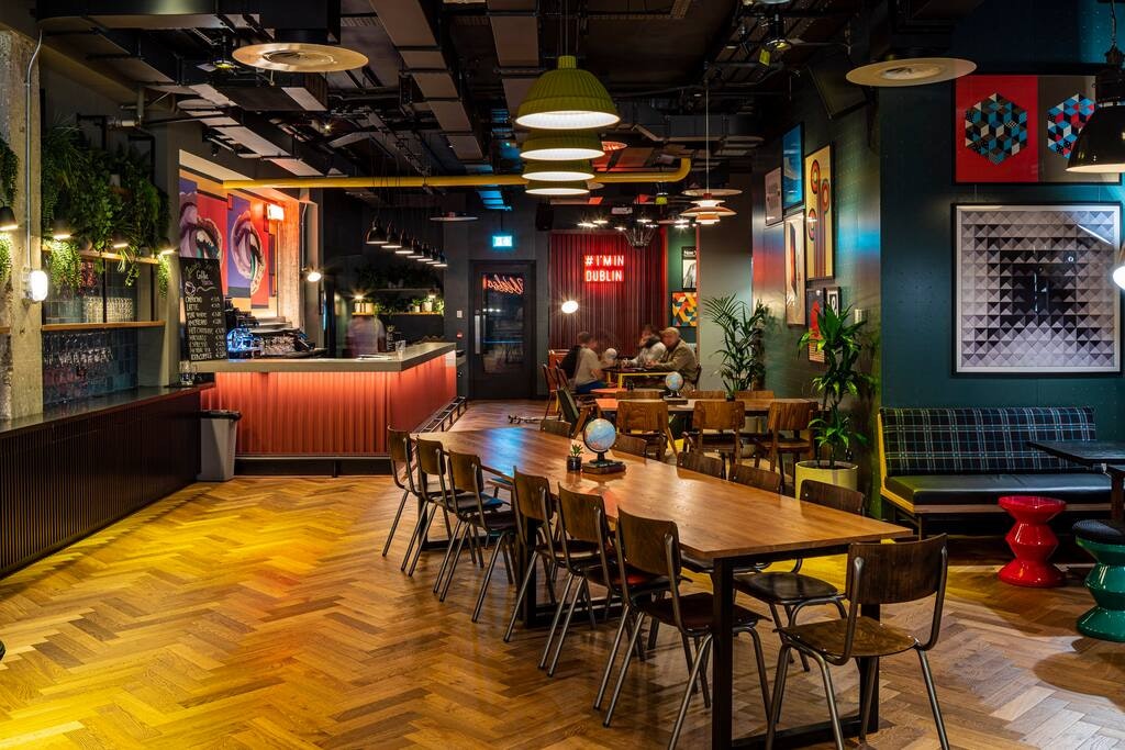 A vibrant dining area features a long wooden table surrounded by wooden chairs. Colorful artwork adorns the walls, while plants add a touch of greenery. Various pendant lights hang from the ceiling, illuminating the space, which includes a bar area in the background.