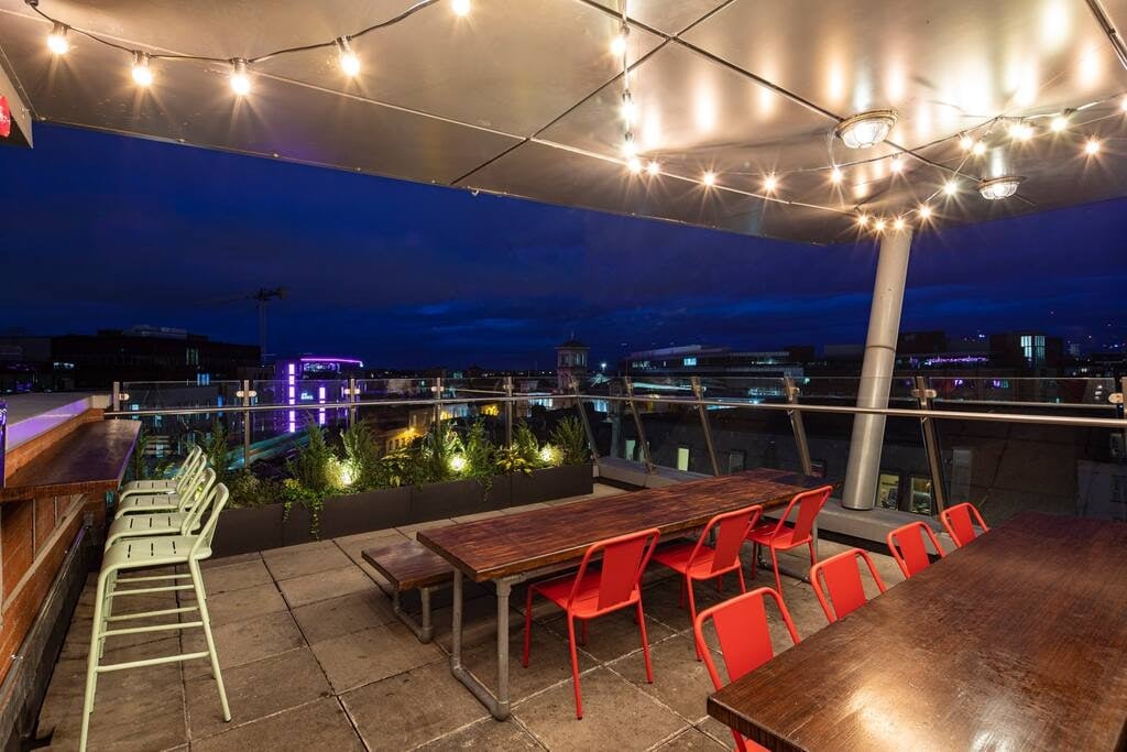 A rooftop terrace is illuminated by string lights, featuring several wooden tables surrounded by red and green chairs. Lush plants are integrated into the decor, adding greenery against the evening sky. A cityscape is visible in the distance, enhancing the setting.