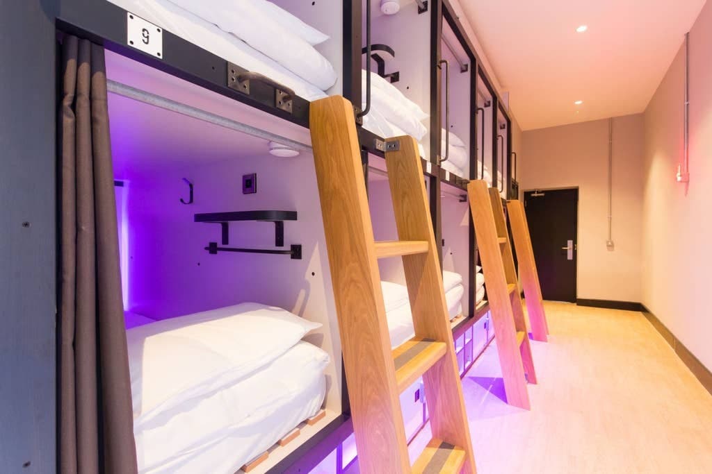 A row of modern sleeping pods is arranged along a well-lit corridor. Each pod features a wooden ladder for access, with neatly made bedding visible inside. Soft purple lighting enhances the space, contributing to a contemporary and organized environment.