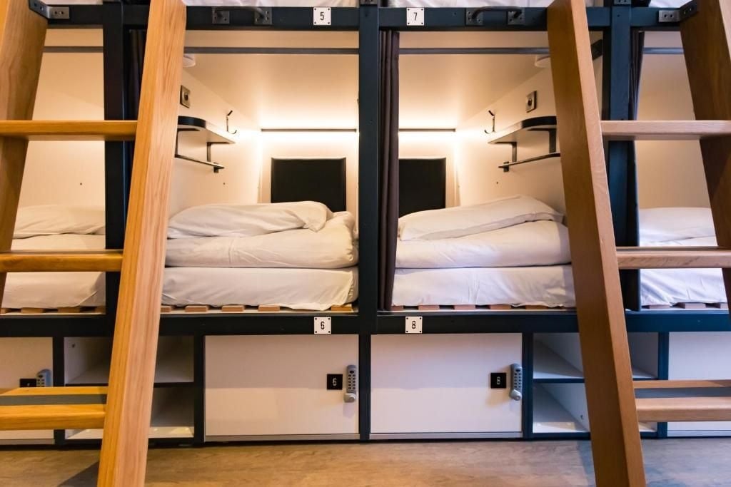 A neatly arranged sleeping area features multiple bunk beds with white bedding. The design includes wooden ladders for access. Each bed is individually positioned within a cubicle-like structure, providing a sense of privacy. Soft lighting enhances the overall ambiance.