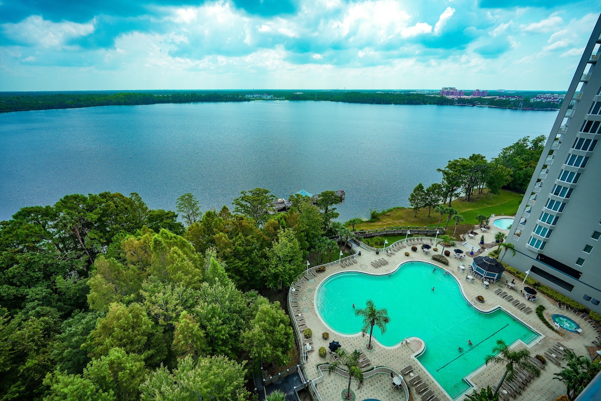 A spacious outdoor pool area is set beside a calm lake, surrounded by lush greenery. Lounge chairs are arranged around the pool, and several guests can be seen enjoying the water. The scene is complemented by a view of distant trees and buildings under a partly cloudy sky.