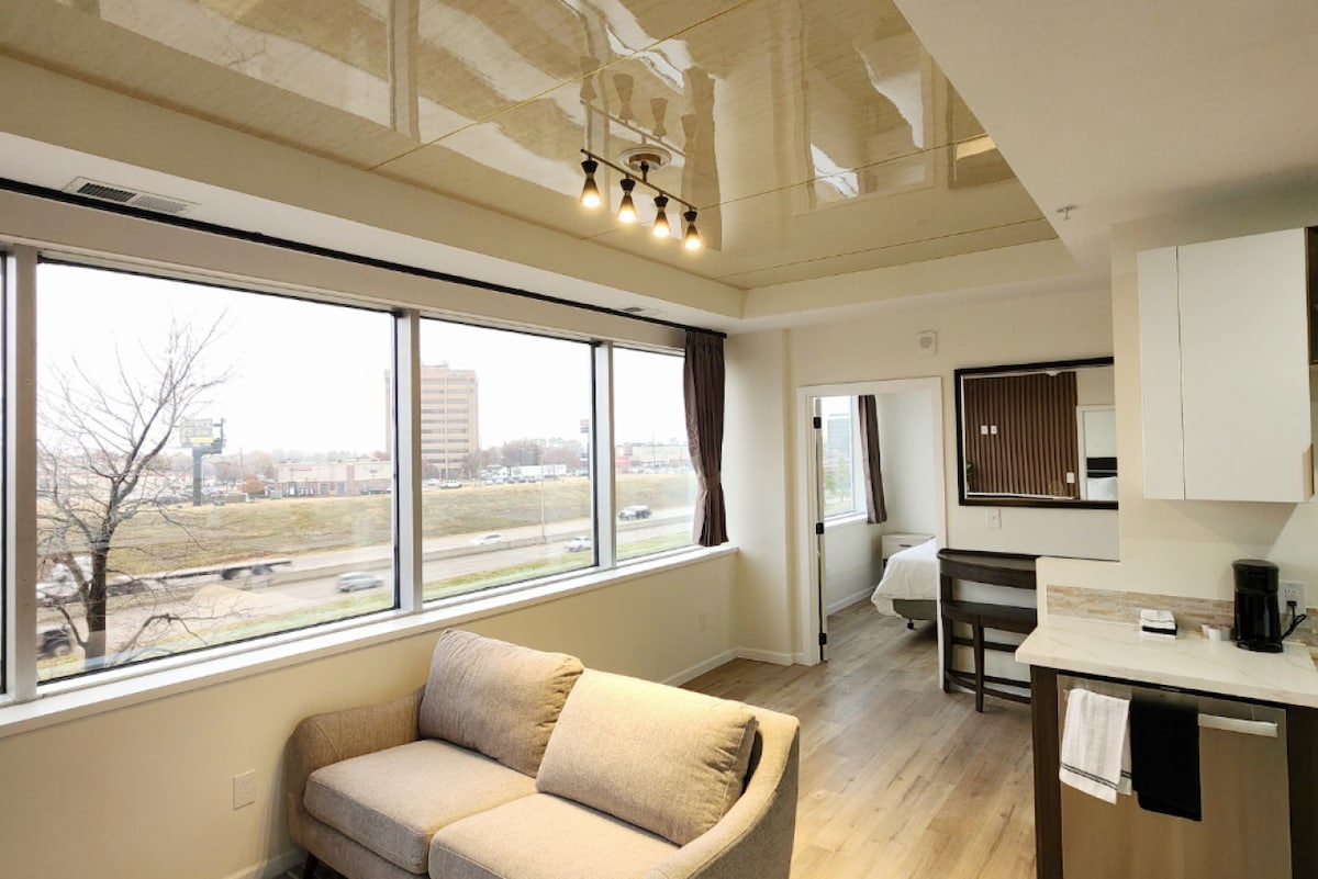 A modern living area features a comfortable gray sofa and a dining table positioned near large windows. Natural light filters through, reflecting off a glossy ceiling. A kitchenette with contemporary cabinetry is located in the corner, enhancing the room's functionality.