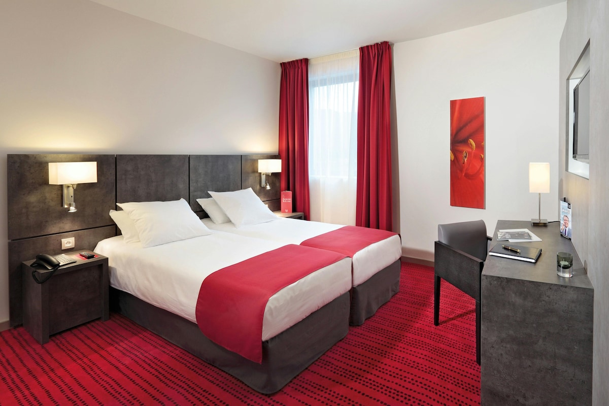 A comfortable studio features two single beds with crisp white linens and decorative red accents. The wall behind the beds showcases modern lighting and artwork. A functional workspace is present, with a chair and desk. Natural light filters in through a bright window, accentuated by rich red curtains.