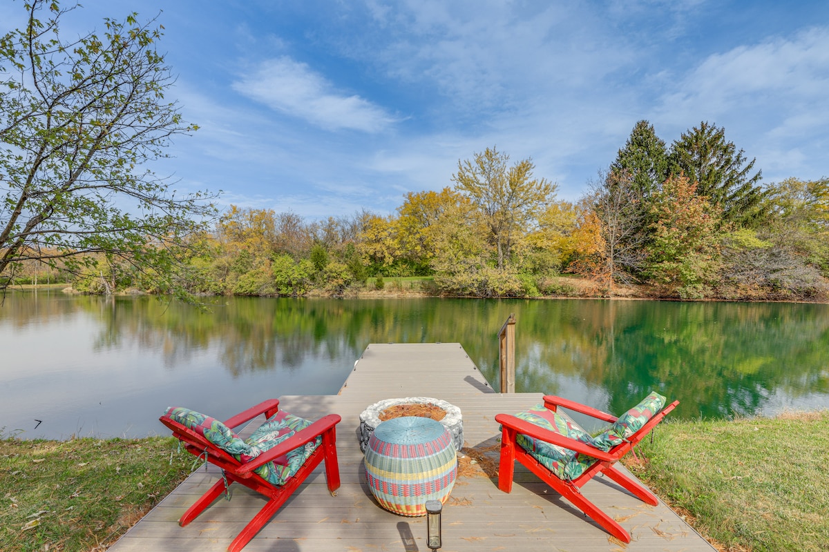 A peaceful fishing dock extends over the calm water, complemented by two red Adirondack chairs facing the serene scene. A decorative barrel serves as a table, while vibrant trees and a clear blue sky enhance the natural setting.