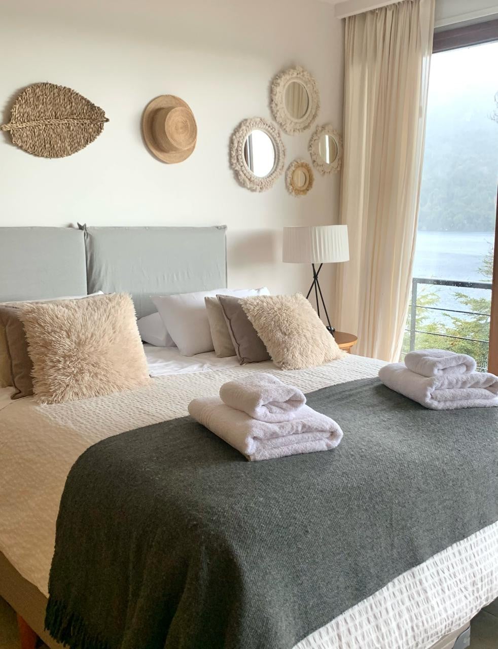 A spacious bedroom features a double bed dressed in soft linens with two rolled towels arranged on top. Natural light enters through a large window, revealing scenic views outside. Various round mirrors and textured wall decor complement the neutral color palette.