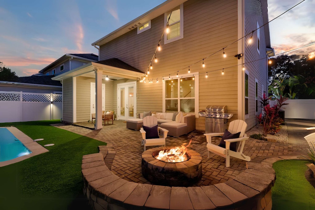 An inviting outdoor space features a comfortable seating arrangement around a fire pit, surrounded by a landscaped patio. A heated saltwater pool reflects the evening sky, while bistro lights create a warm glow over the area, enhancing the inviting atmosphere for relaxation.