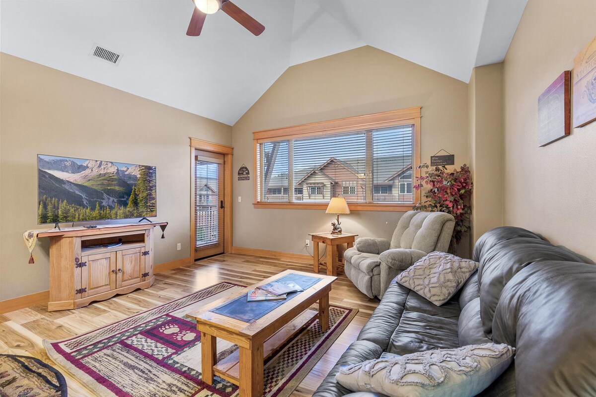The open-concept living area features a comfortable leather sofa and a cozy armchair. A wooden coffee table sits on a patterned area rug. A flat-screen TV is positioned on a rustic console. Natural light streams through large windows, providing views of the surrounding area.