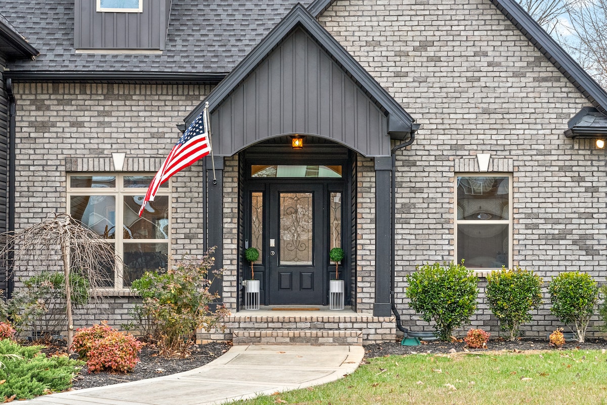 The entrance of the home features a covered porch with dark front doors, framed by elegant glass panels. Flanking the doors are decorative potted plants. A flag pole displays the American flag, while neatly trimmed shrubs and seasonal landscaping provide a warm welcome.
