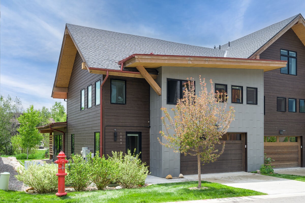 The exterior of a modern mountain home is shown, featuring a combination of dark wood and light grey siding. A well-maintained lawn surrounds the property, with flowering shrubs and small trees enhancing the landscape. A driveway leads to the garage, and a fire hydrant is visible nearby.