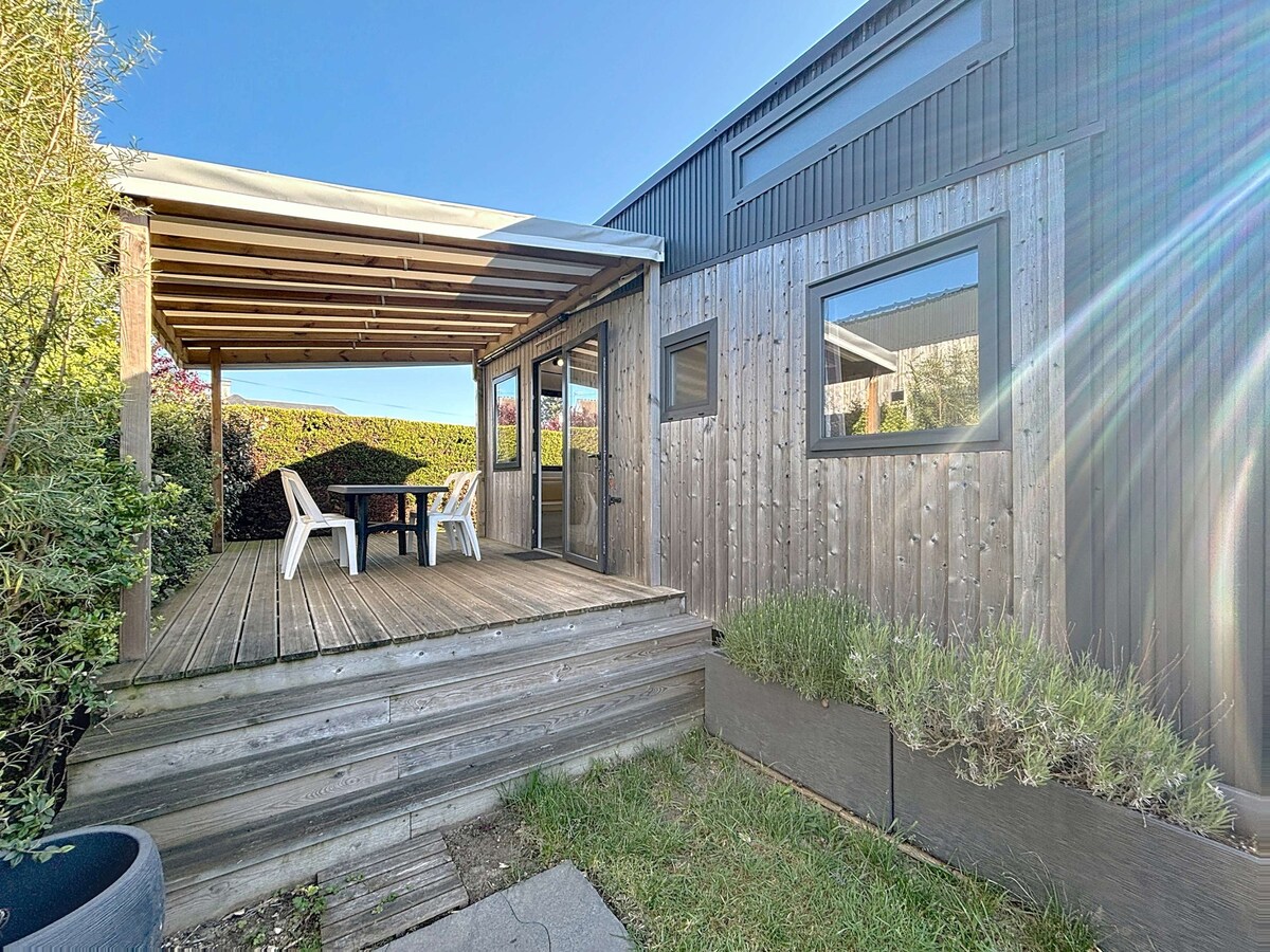 A wooden terrace is featured at the entrance of the Tiny House, with a table and two chairs positioned for outdoor dining. Lush greenery surrounds the area, and large windows provide natural light to the interior. The structure is complemented by a metal roof.