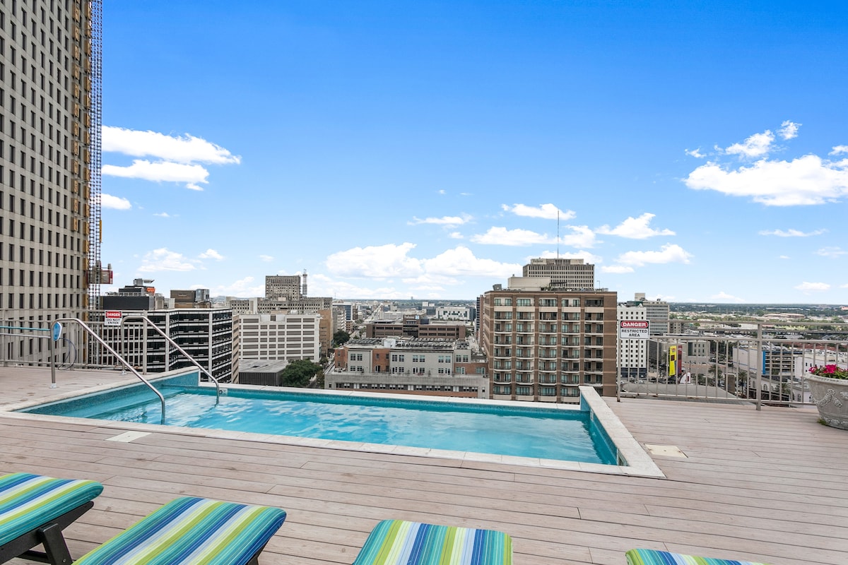 A rooftop pool area is visible, offering scenic views of the downtown skyline. Lounge chairs with striped cushions are arranged around the pool deck, providing space for relaxation. The clear blue water of the pool contrasts with the wooden deck, under a clear sky with scattered clouds.