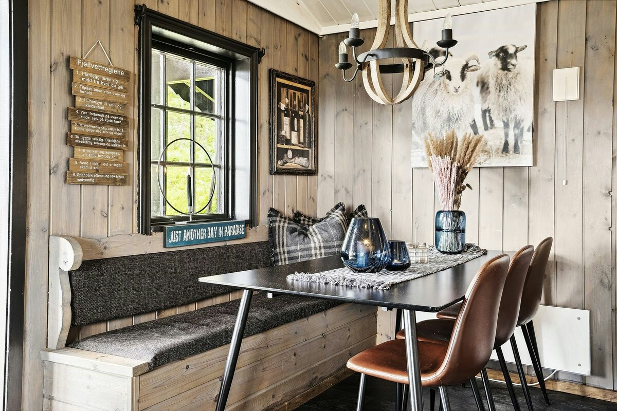 A cozy dining area features a wooden table surrounded by four chairs. A built-in bench provides additional seating against the wall. Decorative elements include a modern chandelier and framed artwork of sheep. Natural light filters through a window, enhancing the inviting space.