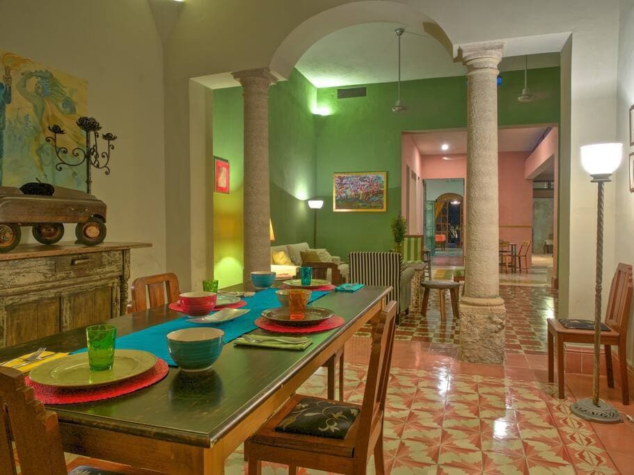 A spacious dining area features a large table set for a meal, decorated with vibrant dishware in blue, green, and terracotta. Tall columns support the ceiling, and colorful walls enhance the inviting ambiance. A view of the adjoining room with artistic decorations is visible in the background.