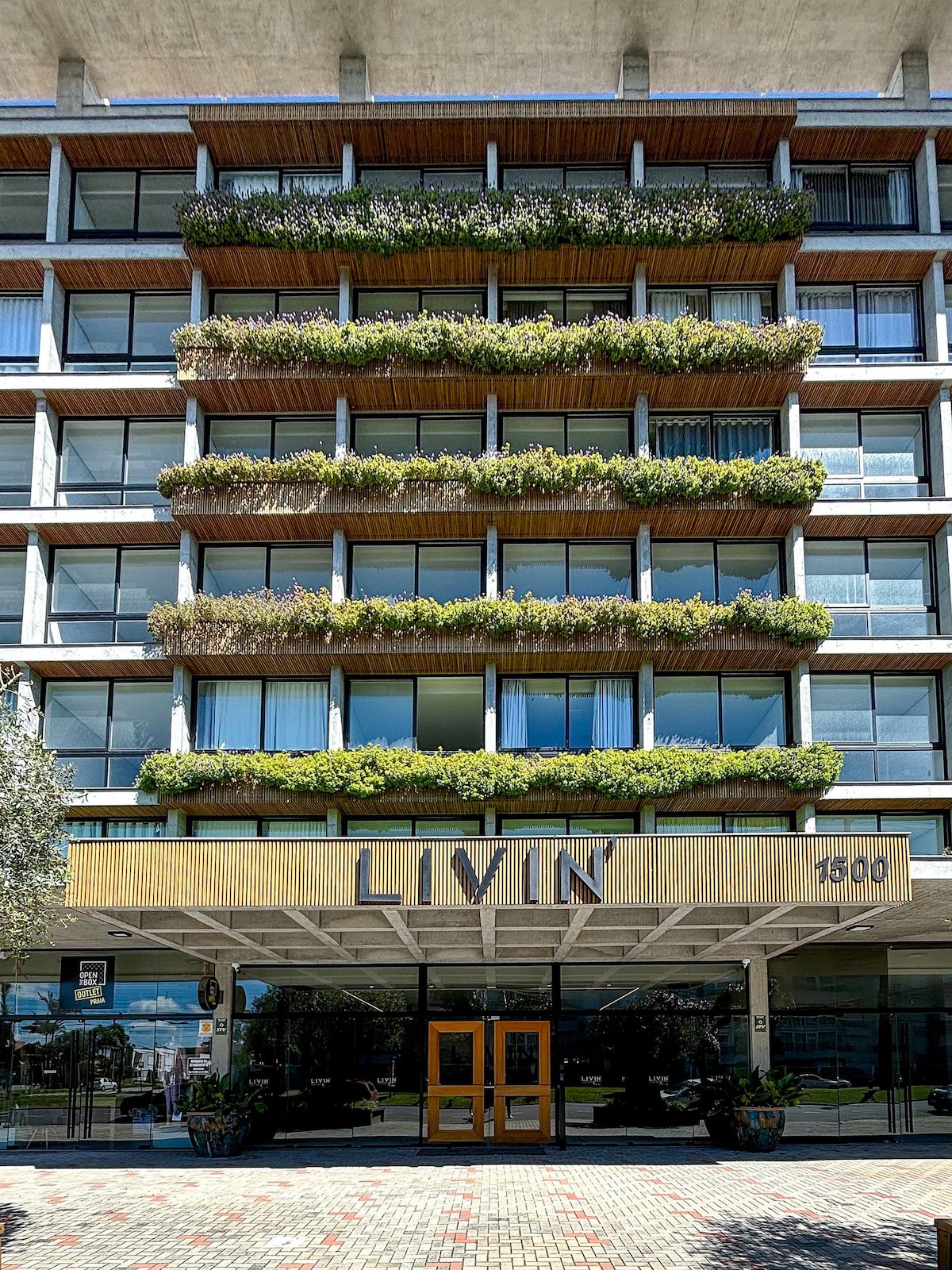 The modern building features a multi-level facade adorned with planters filled with greenery. Large glass windows provide a view into the welcoming space within. The entrance is marked by wooden double doors, flanked by neatly arranged stones on the ground.