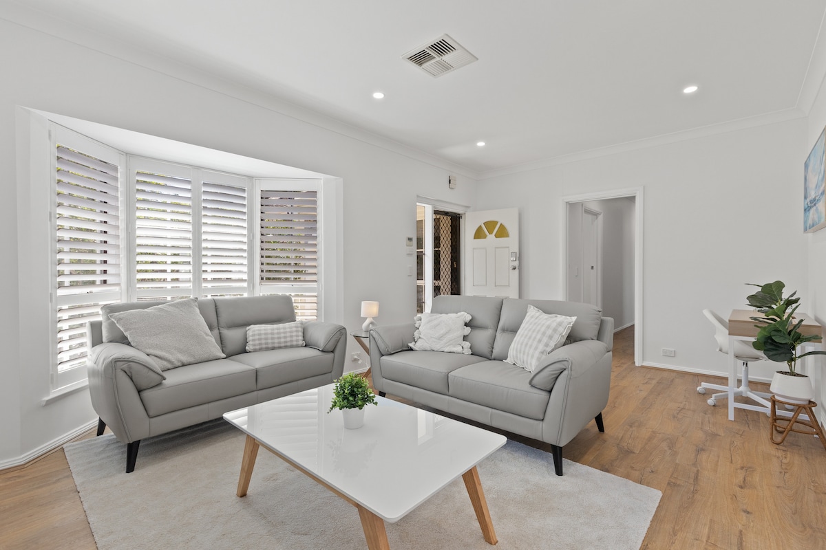 A bright living room showcases two light gray couches and a modern coffee table atop a soft area rug. Natural light enters through window shutters, illuminating the spacious layout. A small study nook with a chair and plant is visible in the background.