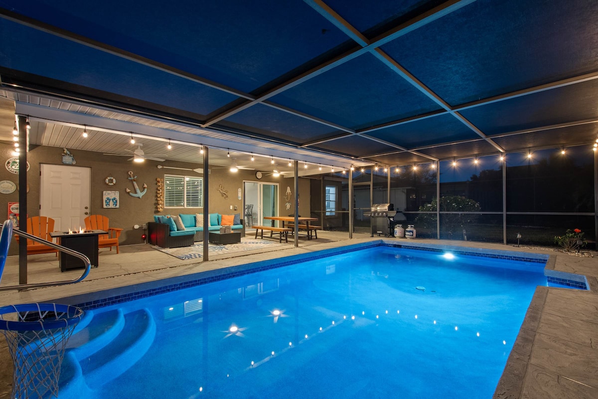 An inviting outdoor space showcases a heated pool surrounded by a screened-in patio. String lights gently illuminate the area, highlighting cozy seating and a dining table, while a BBQ grill is positioned conveniently for outdoor meals.