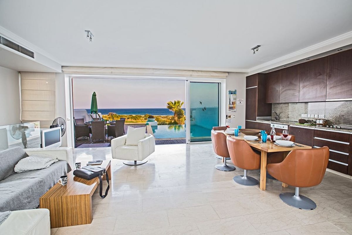 An open living area features a comfortable gray sofa, a wooden coffee table, and a stylish round dining table with orange chairs. Large glass doors provide a view of the sea and lead to the outdoor space, enhancing natural light and inviting coastal scenery.