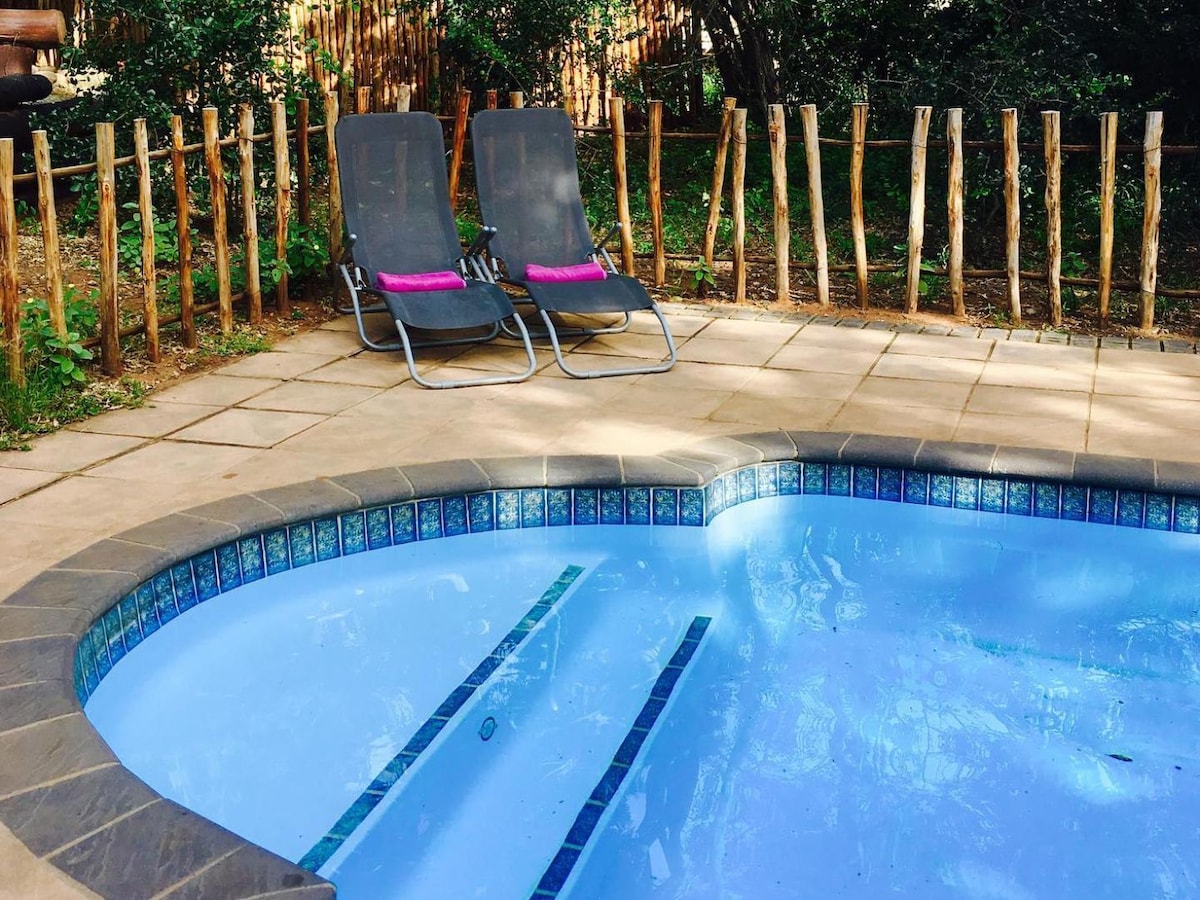 A small outdoor pool is surrounded by natural stone tiles. Two lounge chairs with cushions are positioned nearby, offering a space to relax. A rustic wooden fence provides a barrier, and greenery from surrounding trees creates a peaceful ambiance.