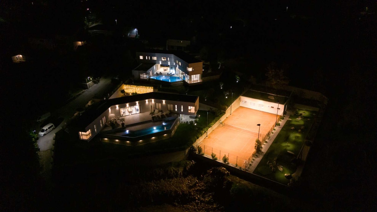 The villa is displayed from an aerial perspective at night, revealing its modern architecture and illuminated outdoor spaces. The private pool and terrace glow softly, while a well-lit tennis court is visible nearby, surrounded by landscaped areas, indicating a serene and inviting retreat.