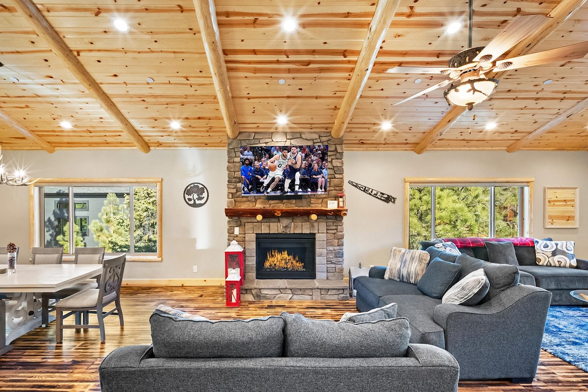 The open living area features vaulted pine ceilings with log accents. A cozy gas fireplace is framed by natural stone, and a large Smart TV is mounted above. Two plush sofas, arranged for relaxation, face toward the inviting fireplace, while a formal dining table is visible in the background.