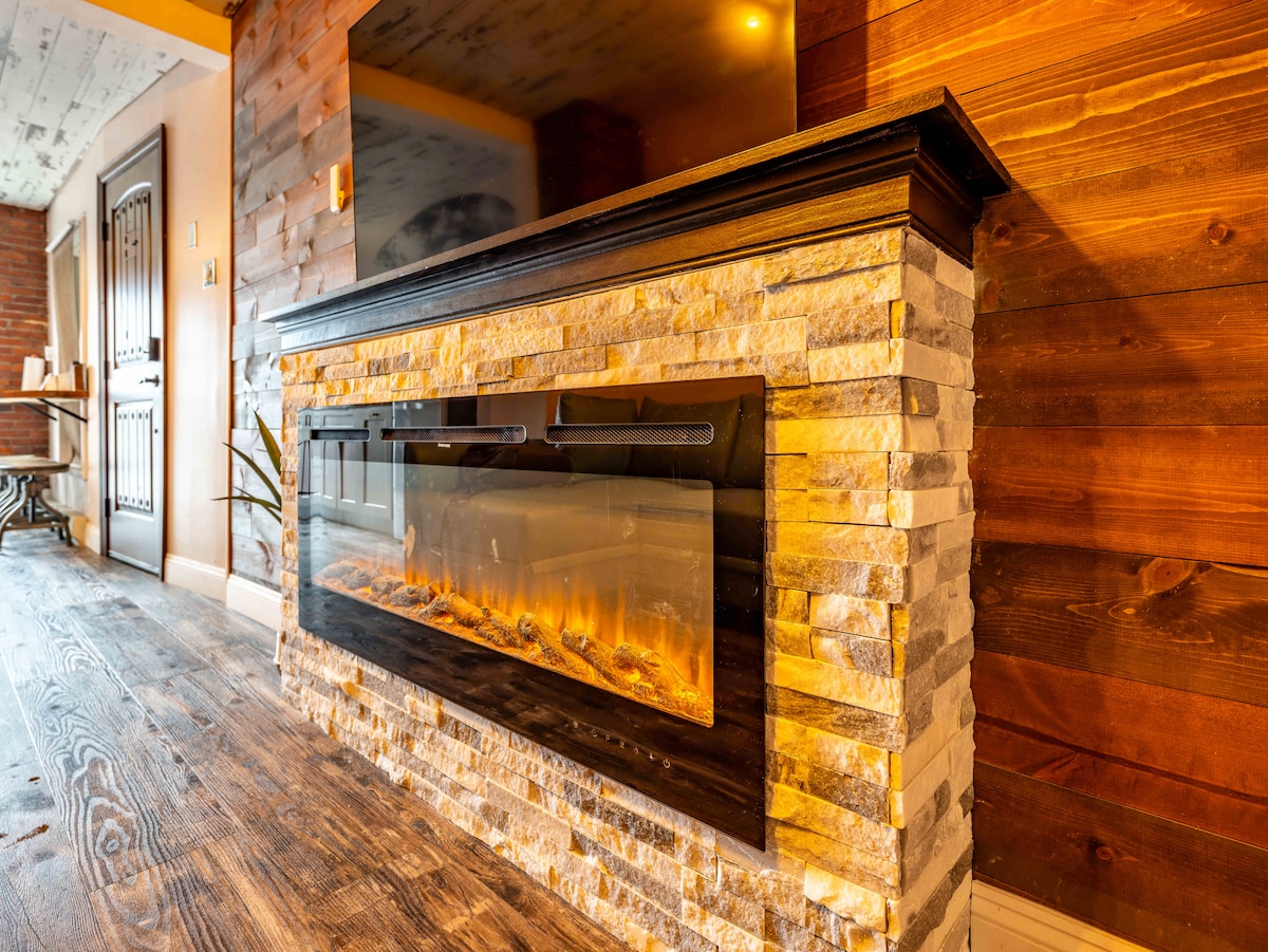 An electric fireplace is set into a stone feature wall, providing a warm focal point in the living area. Natural wood paneling adds texture, while light reflects gently off the surface. The sleek design complements the contemporary aesthetic of the space.