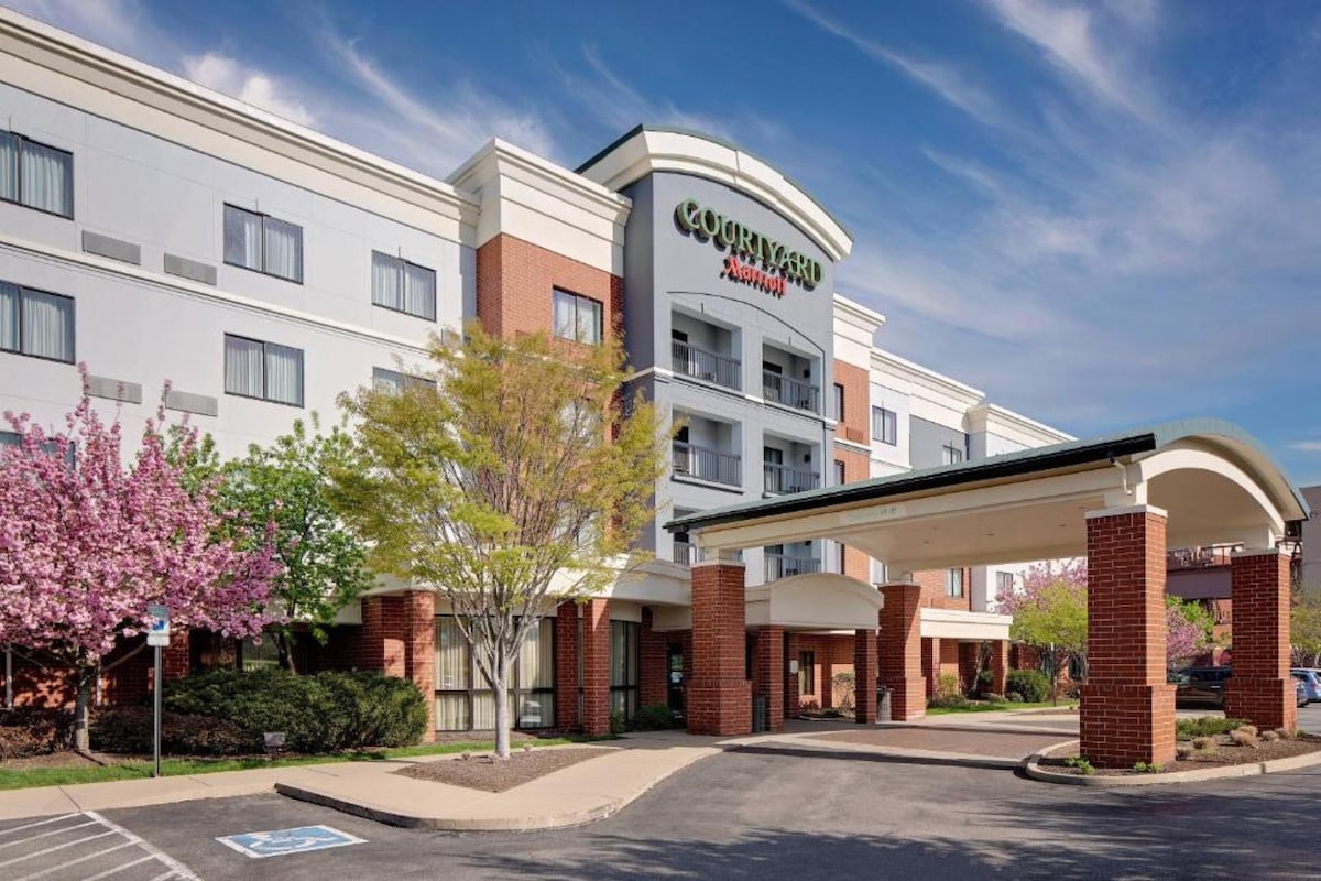 The exterior of the Courtyard Pittsburgh Waterfront is shown, featuring a modern three-story building with a welcoming entrance. Lush green landscaping and blooming trees enhance the appeal. A blue sky with scattered clouds creates a bright and inviting atmosphere.