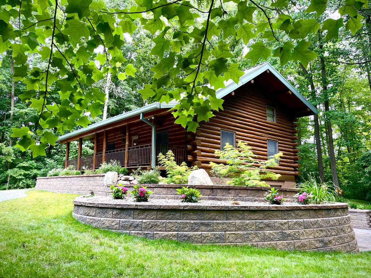 A spacious log cabin rests in a wooded setting, accented by soft landscaping. Flowering plants enhance the front area, while the cabin's broad porch provides a welcoming entrance. Tall trees surround the property, contributing to a sense of tranquility and seclusion.