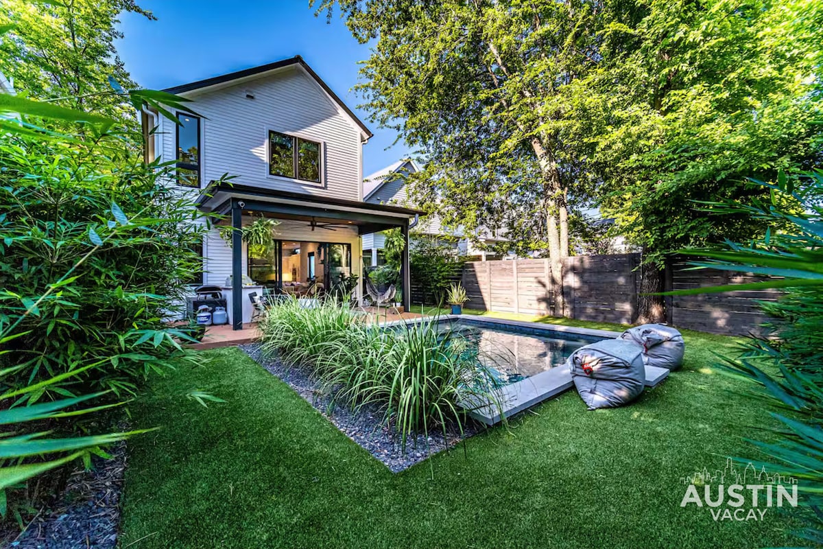 A modern home is presented from the backyard, showcasing a lush green lawn surrounding a sparkling plunge pool. A shaded patio with seating provides a relaxing area, while surrounding trees contribute to a serene atmosphere in this inviting outdoor space.