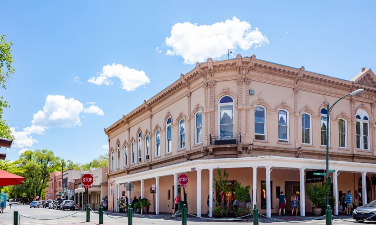 A classic building with intricate architectural details is featured in the image. Large arched windows reflect the sky, which is adorned with fluffy white clouds. Street signs indicate pedestrian areas, while people are seen walking and enjoying the vibrant surroundings.