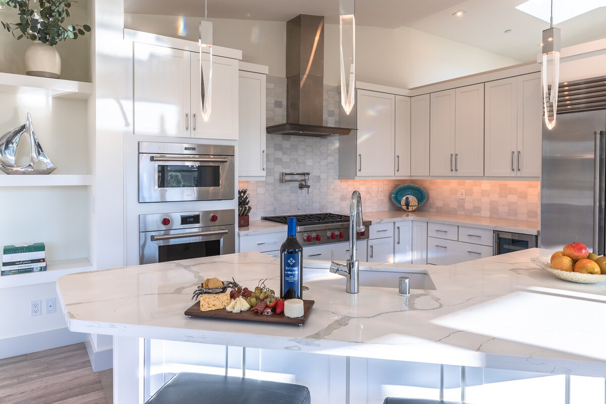 The kitchen features a large, central island with a marble countertop, complemented by state-of-the-art stainless steel appliances. A platter of cheese, fruit, and a bottle of wine is arranged on the island, while pendant lights add illumination above the space.