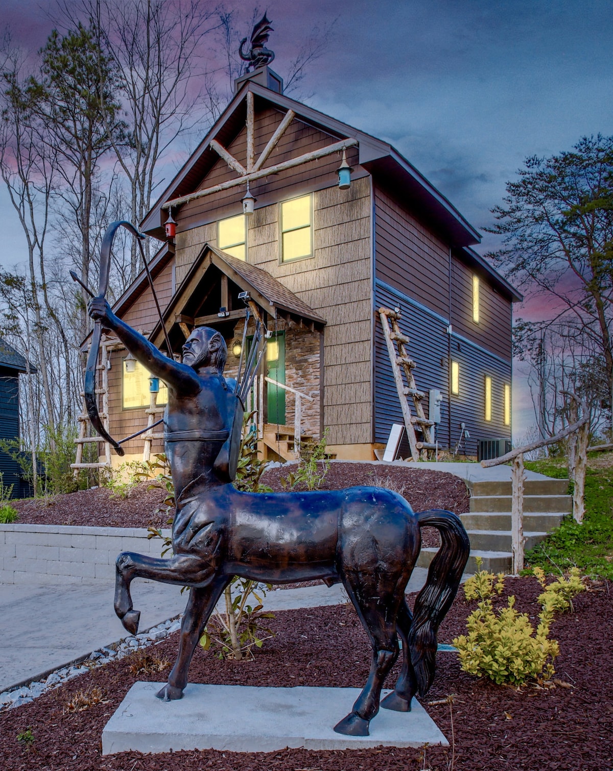 A centaur statue stands in front of a multi-level structure, showcasing a rustic design with wooden siding and large windows. The house features a steeply pitched roof, adorned with colorful accents. Surrounding gardens include newly planted greenery and soft landscaping, set against a twilight sky.