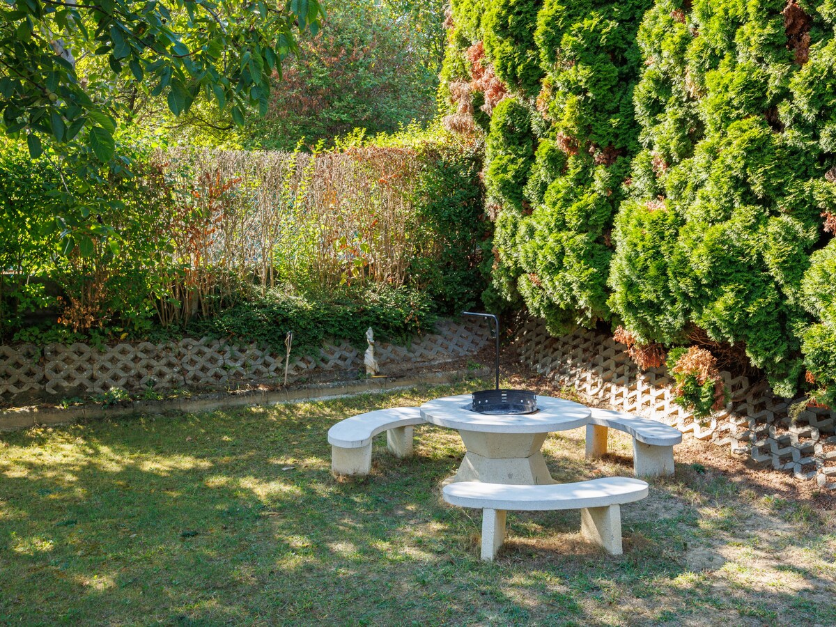 An outdoor area is depicted, featuring a circular stone table surrounded by curved stone benches. Lush greenery, including tall hedges, frames the space, creating a serene environment. The ground is covered in grass, with a small decorative feature visible in the background.