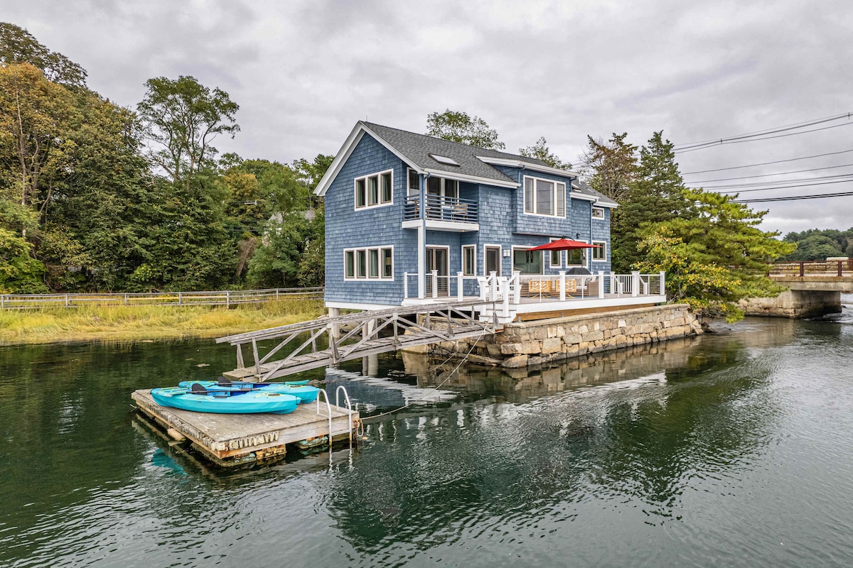 A charming blue house is positioned over water, featuring multiple outdoor spaces. An expansive deck is visible with a red umbrella and seating, while two kayaks rest on a nearby floating dock. Surrounding greenery enhances the natural setting.