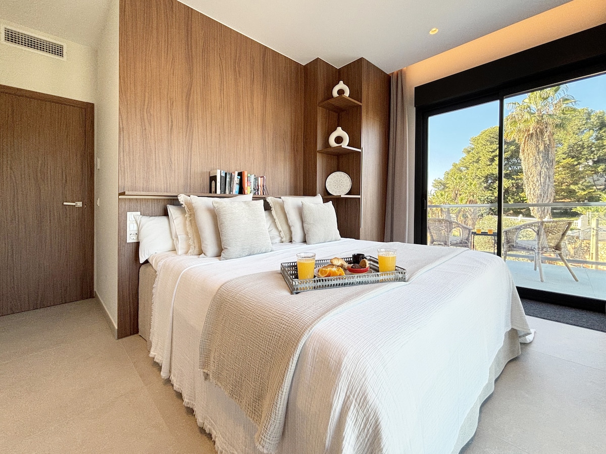 Acogedor dormitorio secundario con una cómoda cama doble y un cabecero de madera de diseño. Incluye almacenamiento incorporado, estanterías decorativas y una amplia puerta corredera que proporciona luz natural y acceso a una terraza privada amueblada.