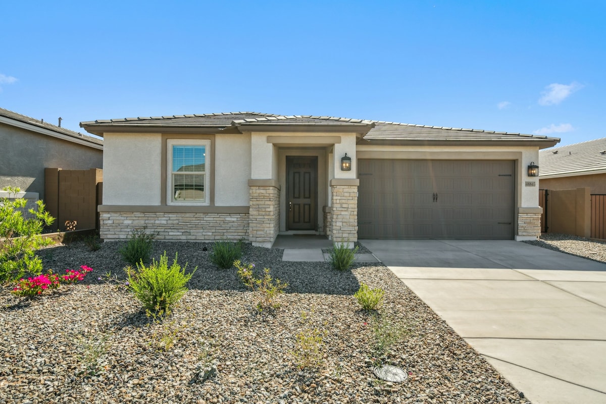 A single-level home is presented featuring a stone-accented exterior and a spacious driveway. The front yard is landscaped with small shrubs and colorful flowers, set against a backdrop of a clear blue sky.