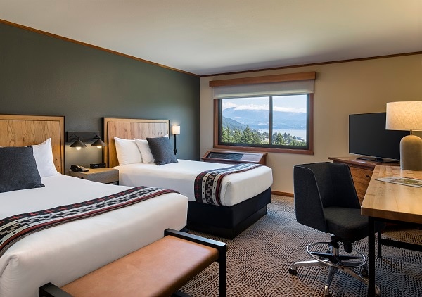 The premier room features two double beds with custom tufted headboards. A large window offers a view of the Columbia River Gorge, flooding the space with natural light. A desk with a lamp, TV, and decorative elements add to the lodge-style decor.