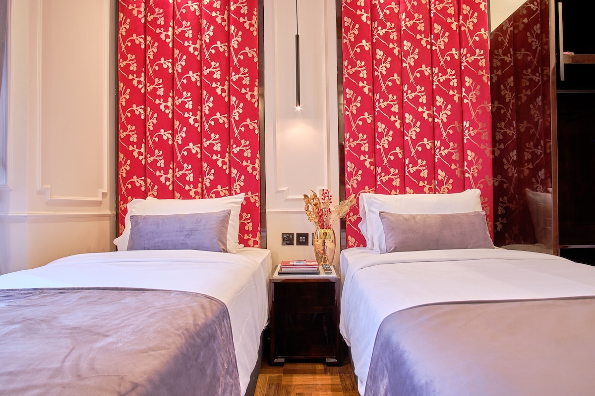 A twin room is presented with two neatly arranged beds adorned with light linens. The headboards feature a bold floral pattern in red, adding a pop of color. A small bedside table holds a decorative vase, contributing to the room's overall elegance.