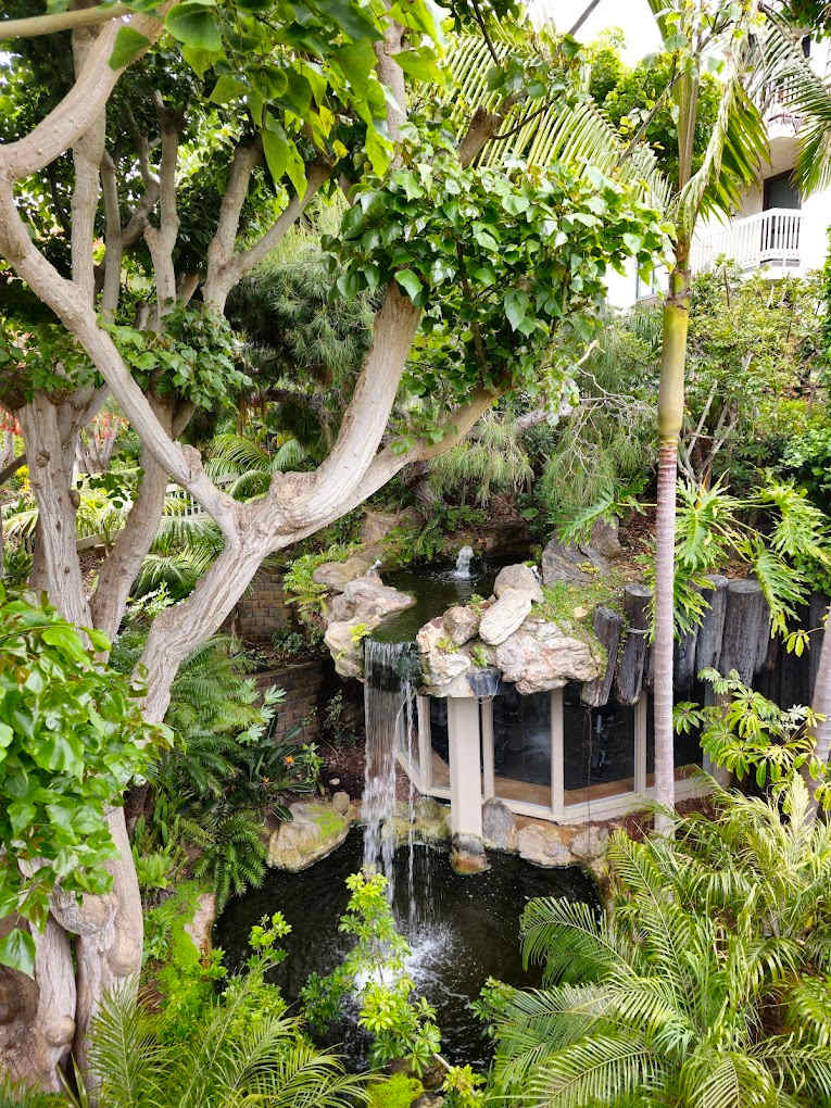 Lush greenery surrounds a serene waterfall cascading into a tranquil pond, framed by tall trees and tropical plants. The natural setting offers a peaceful retreat, with layered textures of rocks and foliage creating a harmonious oasis.