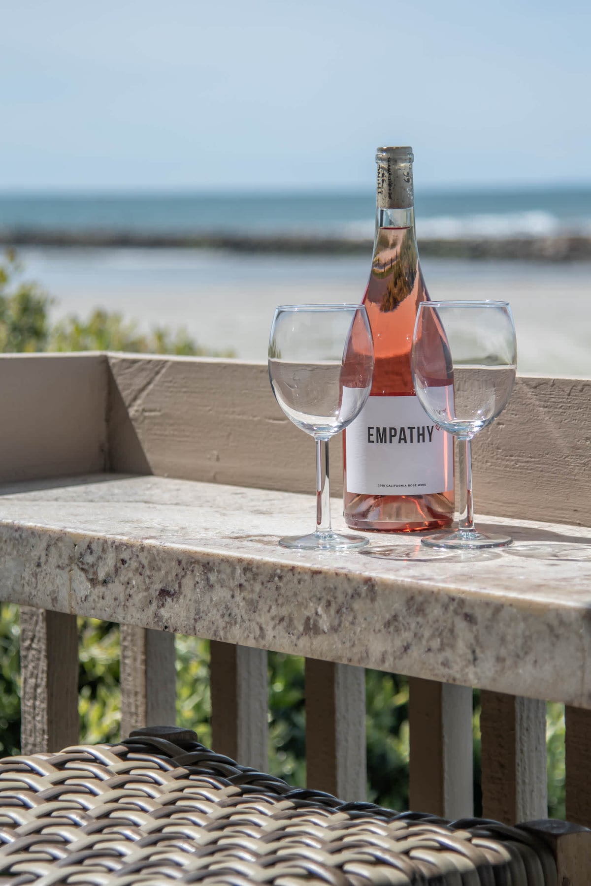 A chic outdoor setting features a bottle of rose wine labeled 'EMPATHY' alongside two clear wine glasses, placed on a textured stone countertop. In the background, the ocean sparkles under a clear blue sky, completing this serene view.