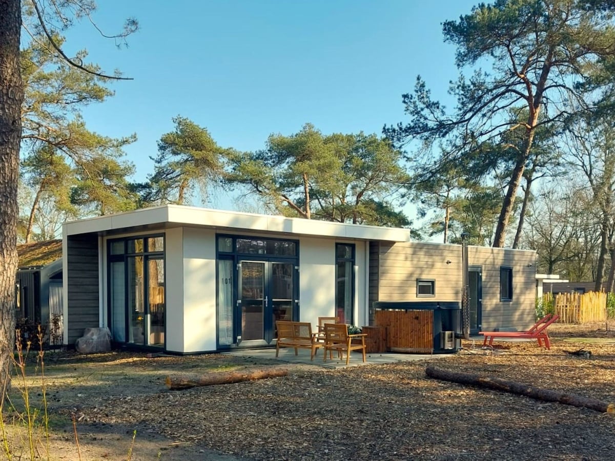 The modern holiday home is surrounded by tall trees, showcasing a blend of wood and glass on its exterior. A private hot tub is visible on the patio, accompanied by a seating area featuring two chairs and a coffee table, ideal for outdoor relaxation.