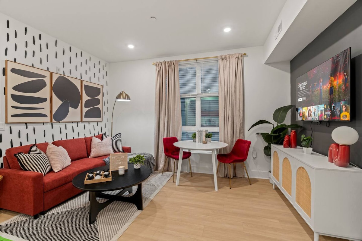A modern living area features a vibrant red sofa paired with two red chairs around a circular table. A large TV is mounted on the wall, displaying a colorful interface. Artistic wall decor and a tall floor lamp add unique style, while light-colored curtains frame the window.