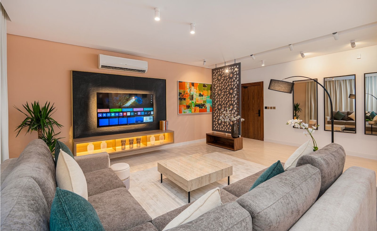 A modern living area is presented, featuring a spacious gray sectional sofa facing a flat-screen smart TV mounted on an accent wall. A decorative divider is positioned to the side, with potted plants and artwork adding color. Soft lighting enhances the inviting space.