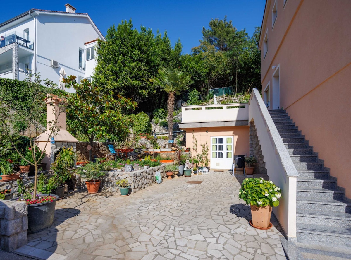 The exterior features a spacious stone courtyard adorned with various potted plants, including flowers and greenery. A flight of stairs leads to an upper level, while a charming outdoor seating area is positioned against a backdrop of lush foliage and a palm tree.