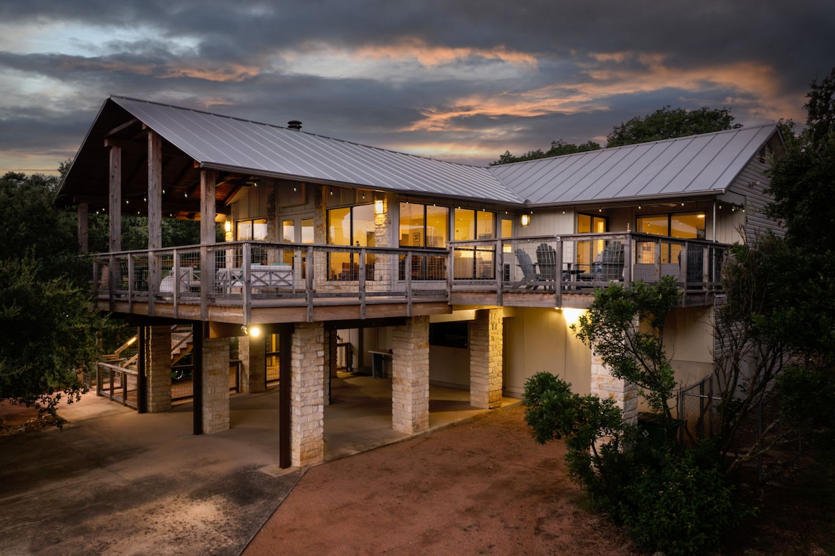 The exterior of a modern home is showcased at dusk, highlighting a wrap-around deck and large windows that illuminate the interior. The structure is elevated on pillars, with stone accents and a metal roof. The surrounding landscape features shrubs and trees.
