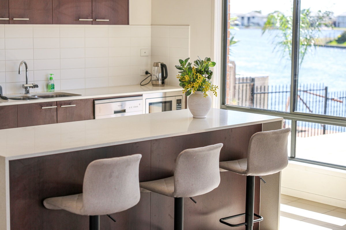 A modern kitchen area features a light-filled counter with three soft upholstered stools. A small white vase with greenery sits on the countertop, complementing the sleek appliances and sink. Large windows offer views of the lake, enhancing the sense of space and light.