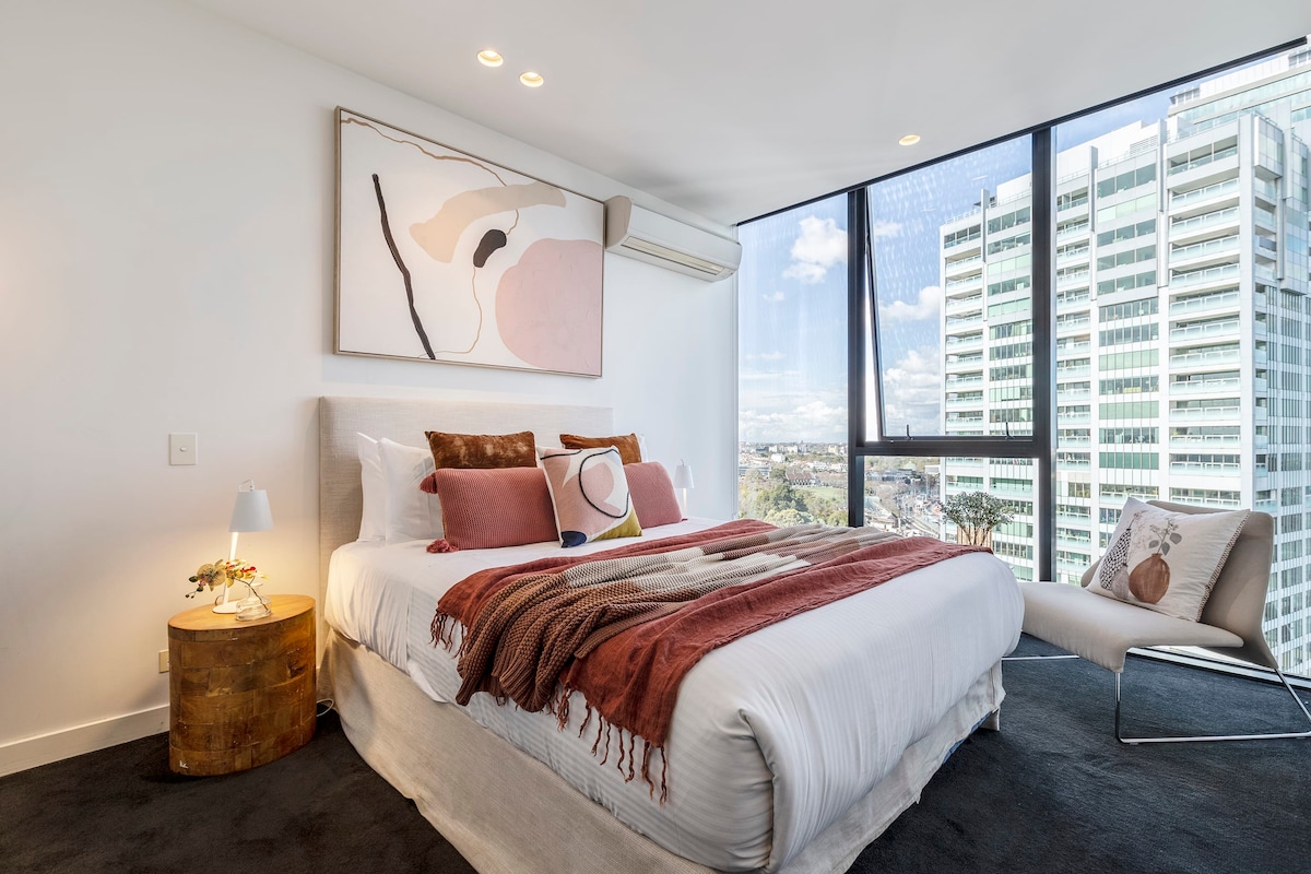 A serene bedroom features a spacious bed dressed in neutral linens and decorative pillows. Natural light floods through large windows, revealing city views. A cozy chair and a small bedside table with a vase of flowers enhance the inviting ambiance.