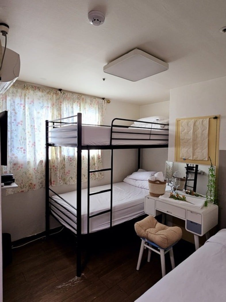 A compact sleeping area is presented, featuring a black metal bunk bed with a twin bed on top and a spacious bed underneath. A small desk with a mirror and decorative plant is located beside the bed, while natural light enters through white floral curtains.