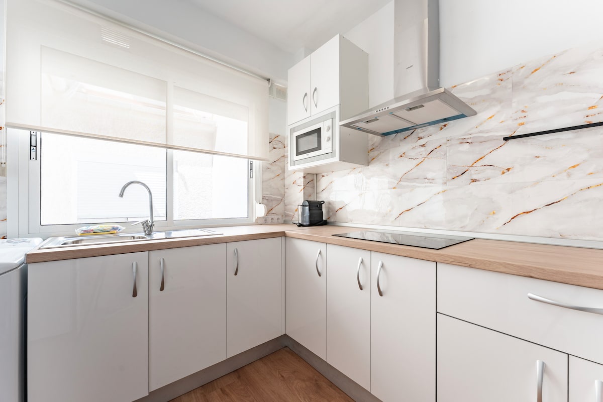 The kitchen features modern cabinetry in white, complemented by light wood surfaces. A large window allows natural light to brighten the space. Essential appliances, such as a microwave and stovetop, are included, along with a sink and ample counter space for meal preparation.