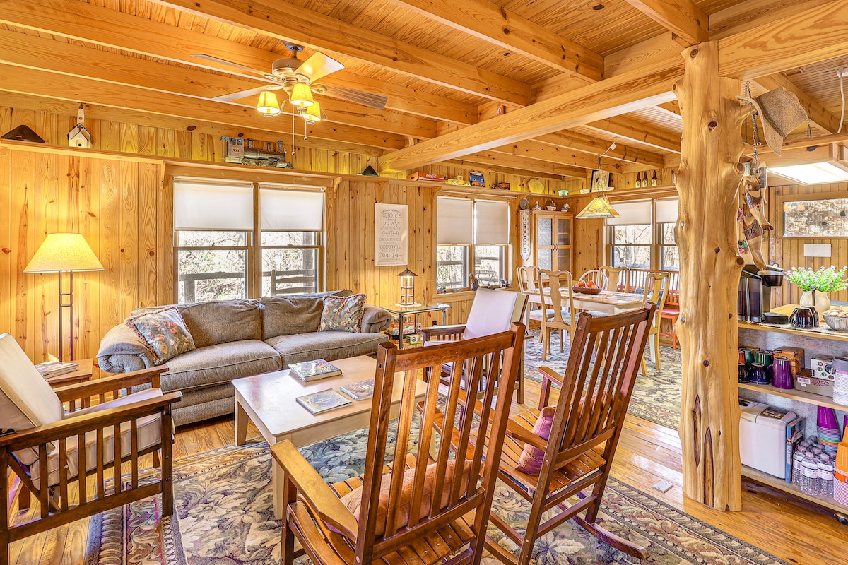 The living area features wooden beams and a warm, rustic decor. A spacious sofa is complemented by several rocking chairs, inviting relaxation. Natural light filters through multiple windows, and a dining table is visible in the background, enhancing the open, welcoming ambiance.